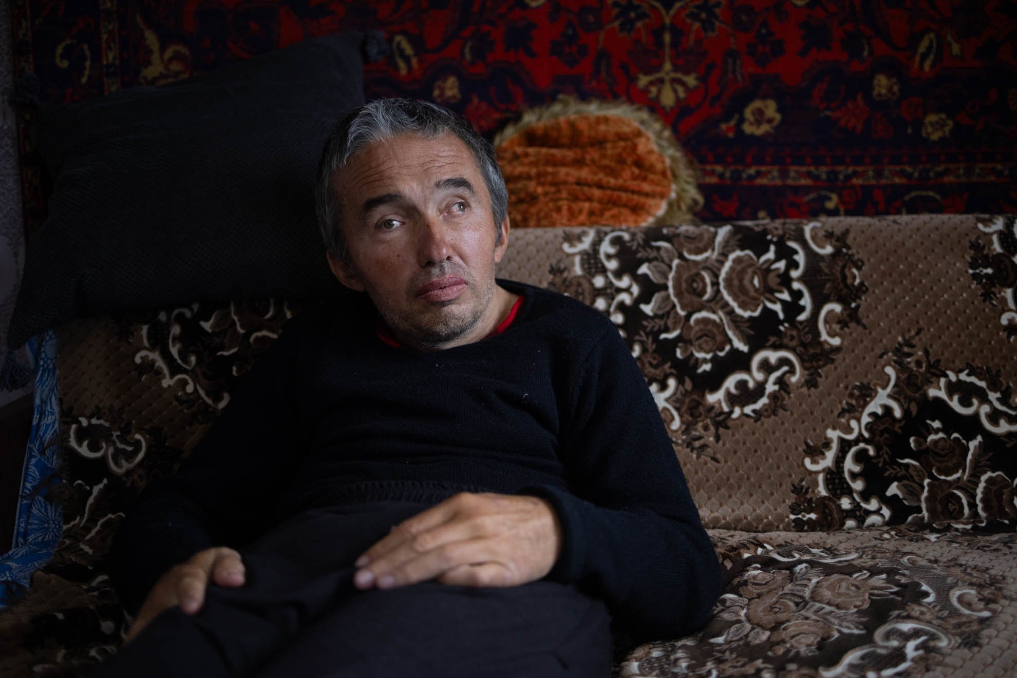 Yevhen is sitting on a brown sofa. He is wearing a black jumper. 