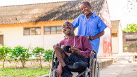 In the foreground, Mario is sitting in his wheelchair and his best friend is behind him, holding the wheelchair handles. The two boys are laughing. In the background, you can make out their school.