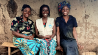 Beatrice, an HI mental health and psychosocial worker and Rose sit on a bench in front of their home. The three women are smiling.