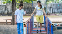 Kanha, an 18-year-old girl learning to walk with an above-the-knee prosthesis on her right leg. She is walking across a wobbly bridge with parallel bars, while an HI rehabilitation staff walks alongside her. They are outside in the yard of the Physical Rehabilitation Centre.